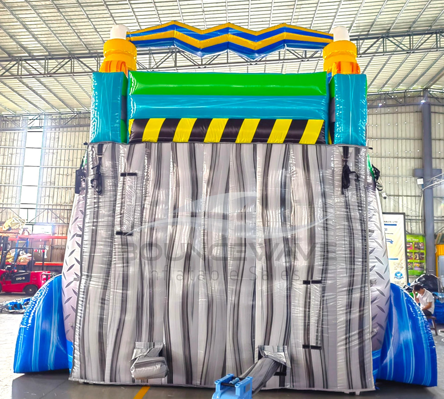 Rear view of 20ft Radioactive Run Hybrid DL water slide showing green and blue color scheme, caution stripes, and enclosed tube slide sections. The slide is set up in an indoor facility with metal roofing and other equipment visible in the background.