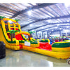 Wide-angle view of 20ft Rasta Splash Hybrid DL water slide featuring yellow, green, and red colors, inflatable palm trees, and a large splash pool area. The slide is set up in an indoor facility with metal roofing, bright lighting, and other inflatable products visible in the background.