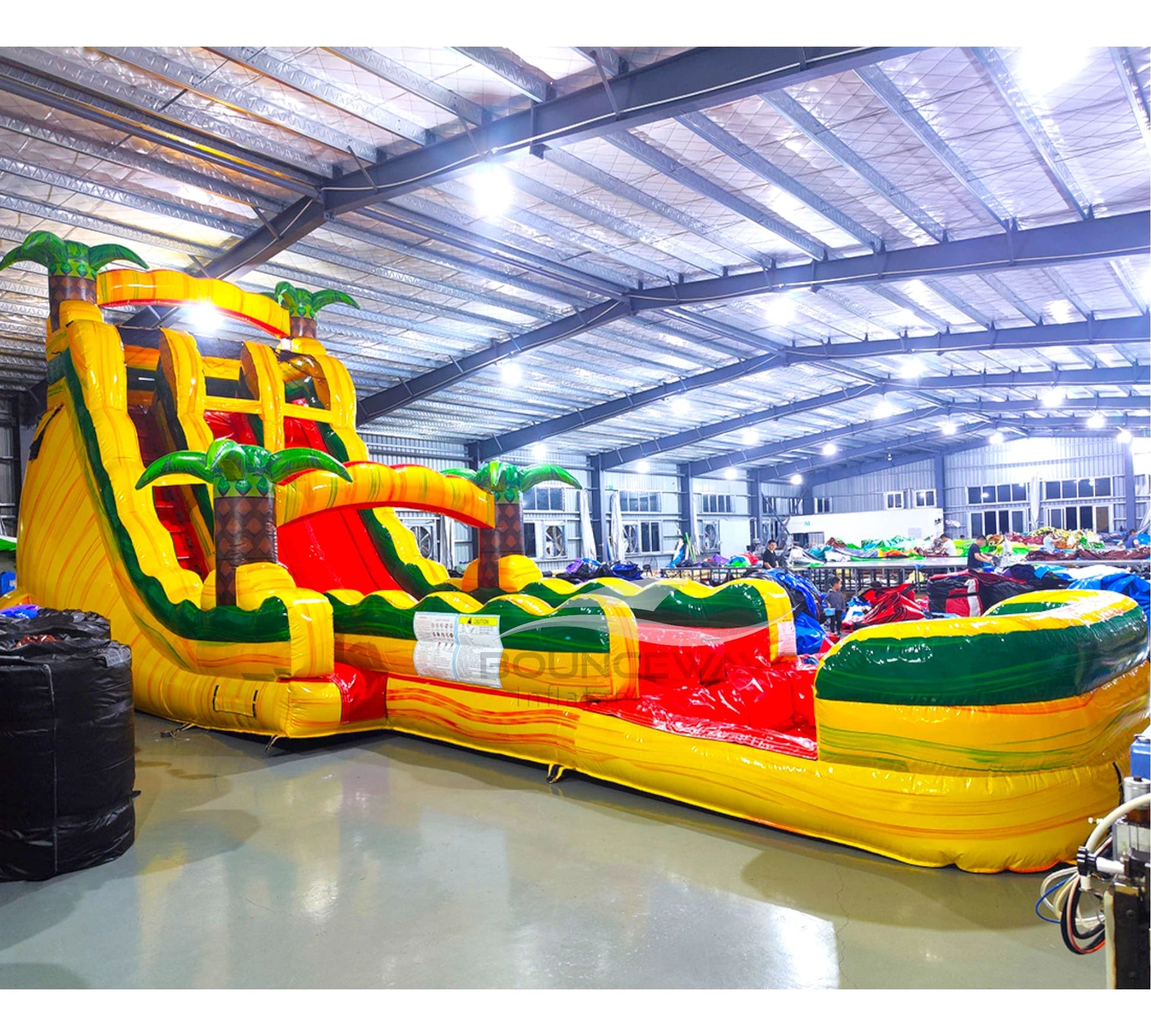 Wide-angle view of 20ft Rasta Splash Hybrid DL water slide featuring yellow, green, and red colors, inflatable palm trees, and a large splash pool area. The slide is set up in an indoor facility with metal roofing, bright lighting, and other inflatable products visible in the background.