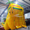 Large inflatable water slide with tropical theme, featuring vibrant yellow, green, and red colors, palm tree decorations, and a spacious splash pool area. The slide combines open and enclosed sections, set up in an indoor warehouse with metal roofing and other equipment visible in the background.