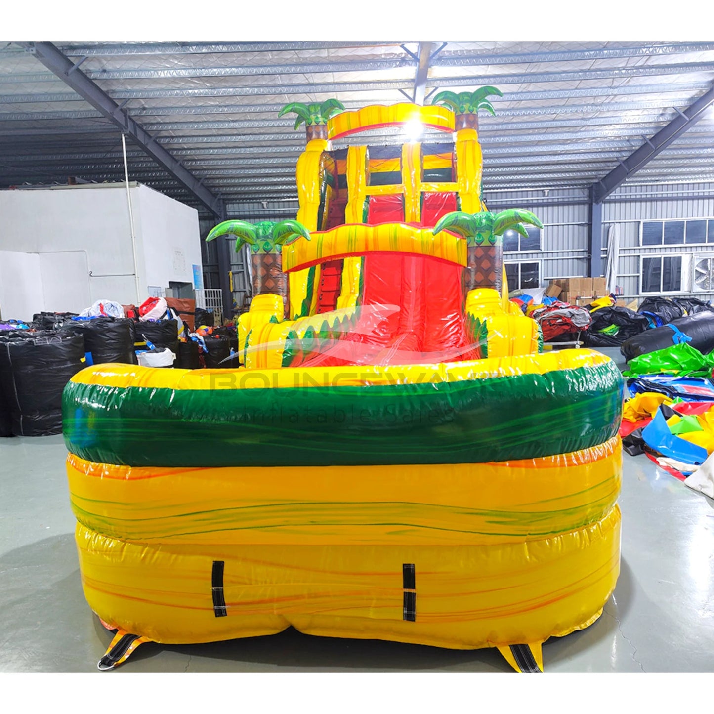 Large inflatable water slide with tropical theme, featuring vibrant yellow, green, and red colors, palm tree decorations, and a spacious splash pool area. The slide combines open and enclosed sections, set up in an indoor warehouse with metal roofing visible.