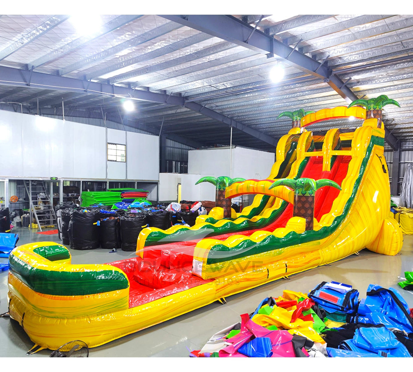 Front angle view of the 20ft Rasta Splash Hybrid DL inflatable water slide, showcasing its yellow, green, and red sliding lanes, palm tree decorations, and spacious splash pool. The slide is set up in an indoor warehouse with other inflatables and equipment visible in the background.