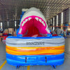 20ft Rip Curl Shark Attack 2pc Hybrid Dual Lane Inflatable Water Slide featuring giant shark mouth entrance, blue water slide, and colorful inflatable base in warehouse
