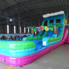 Large inflatable water slide with tropical theme, featuring vibrant blue, green, and pink colors, palm tree decorations, and a spacious splash pool area. The slide has a wave-shaped top and combines open and enclosed sections, set up in an indoor warehouse with metal roofing visible.