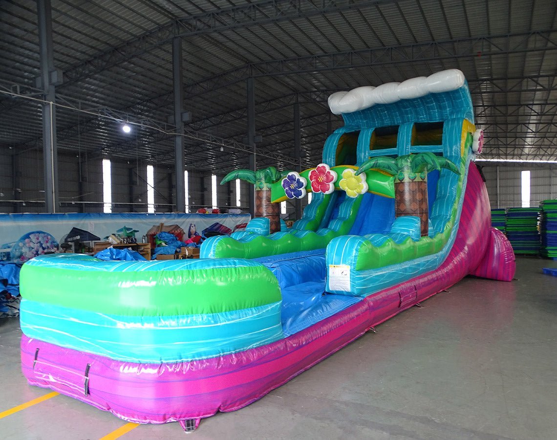 Large inflatable water slide with tropical theme, featuring vibrant blue, green, and pink colors, palm tree decorations, and a spacious splash pool area. The slide has a wave-shaped top and combines open and enclosed sections, set up in an indoor warehouse with metal roofing visible.