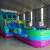 Front angle view of the 20ft Summer Luau Hybrid DL inflatable water slide, showcasing its blue, green, and pink sliding lanes, palm tree decorations, and spacious splash pool. The slide is set up in an indoor warehouse with other inflatables and equipment visible in the background.