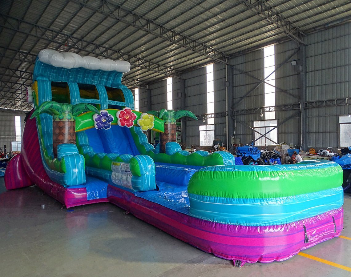 Front angle view of the 20ft Summer Luau Hybrid DL inflatable water slide, showcasing its blue, green, and pink sliding lanes, palm tree decorations, and spacious splash pool. The slide is set up in an indoor warehouse with other inflatables and equipment visible in the background.