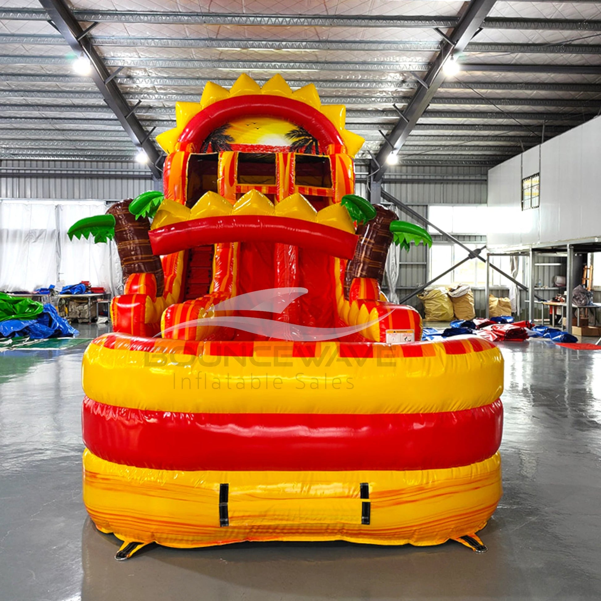 Large inflatable water slide with tropical theme, featuring vibrant yellow and red colors, palm tree decorations, and a sun-shaped top. The slide has multiple lanes, a spacious splash pool area, and combines open and enclosed sections, set up in an indoor warehouse with metal roofing visible.