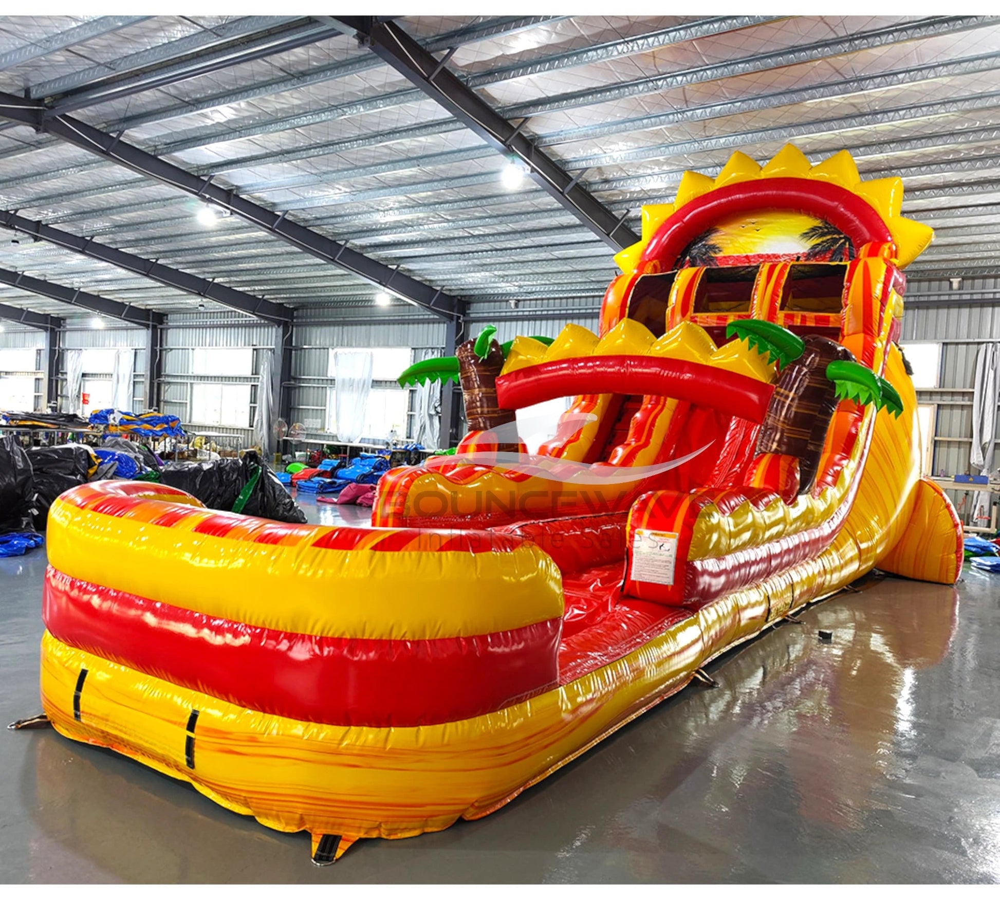 Wide-angle view of 20ft Summer Sizzler Hybrid DL water slide featuring yellow and red colors, inflatable palm trees, sun-shaped top, and a large splash pool area. The slide is set up in an indoor facility with metal roofing, bright lighting, and other inflatable products visible in the background.