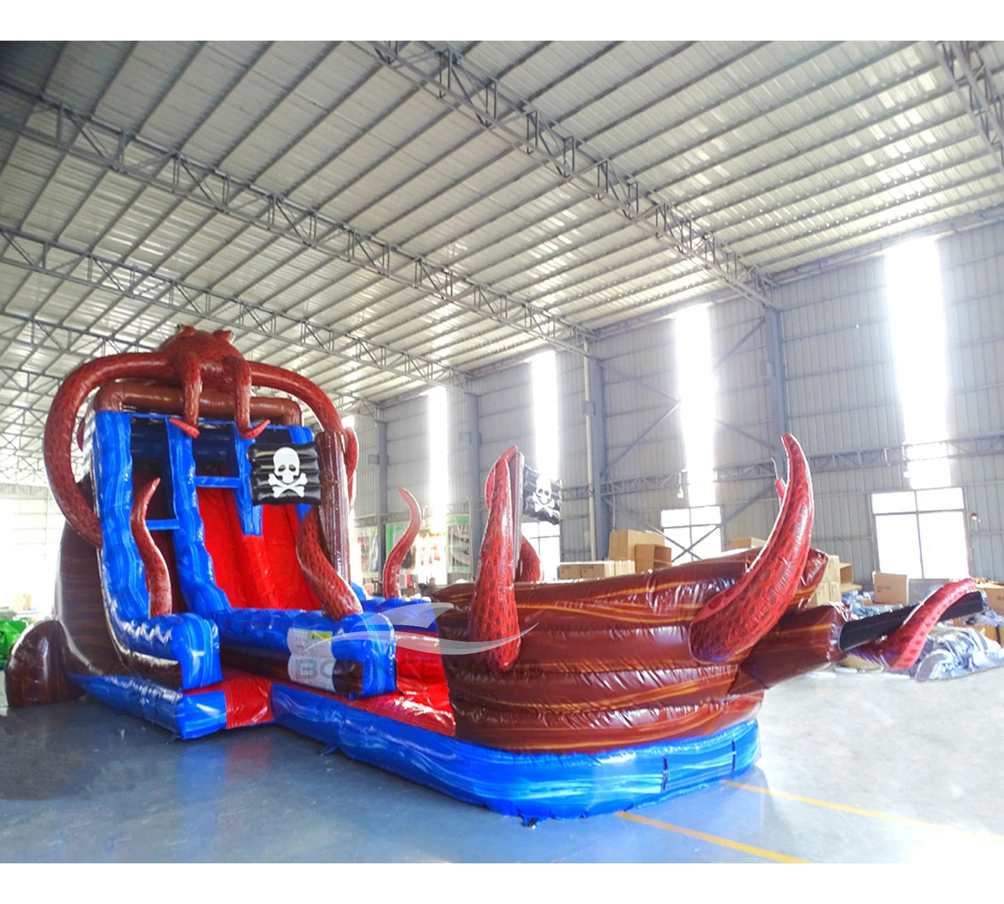 Wide-angle view of 20ft Tentacle Typhoon Hybrid DL water slide featuring red, brown, and blue colors, with prominent octopus tentacles and pirate ship elements. The slide is set up in an indoor warehouse with metal roofing, bright lighting, and other inflatable products visible in the background.