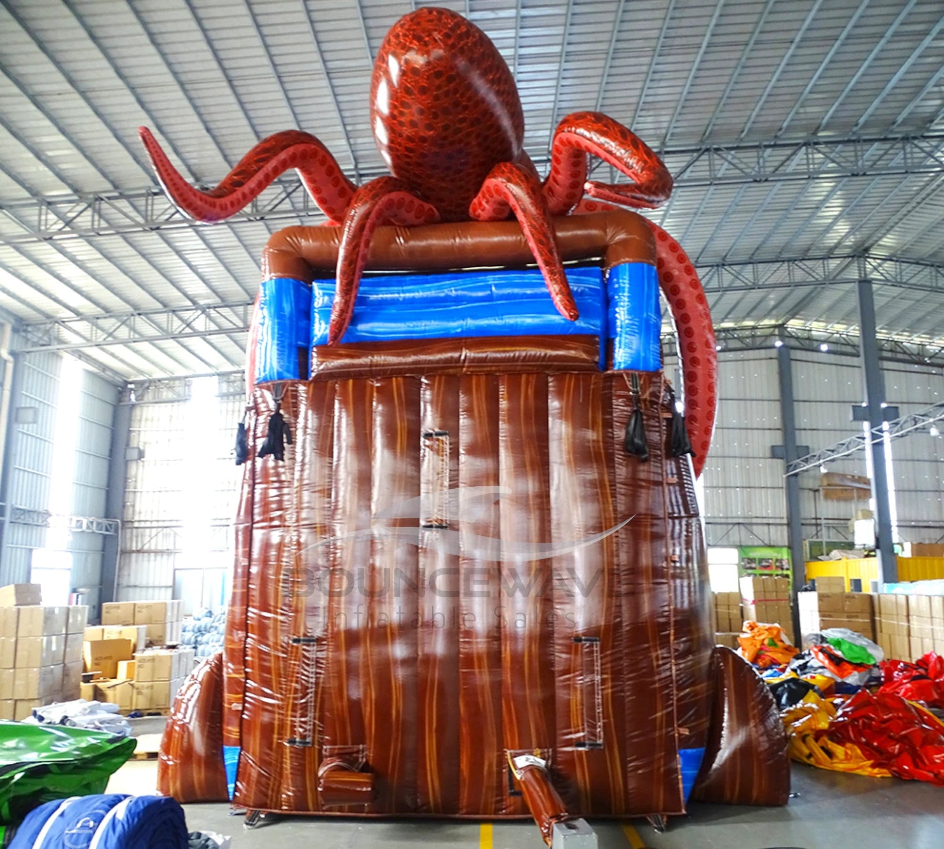 Rear view of 20ft Tentacle Typhoon Hybrid DL water slide showing red, brown, and blue design, with large octopus tentacles wrapping around the structure. The slide features both open and enclosed sliding sections, displayed in an indoor facility with other inflatables and equipment visible in the background.