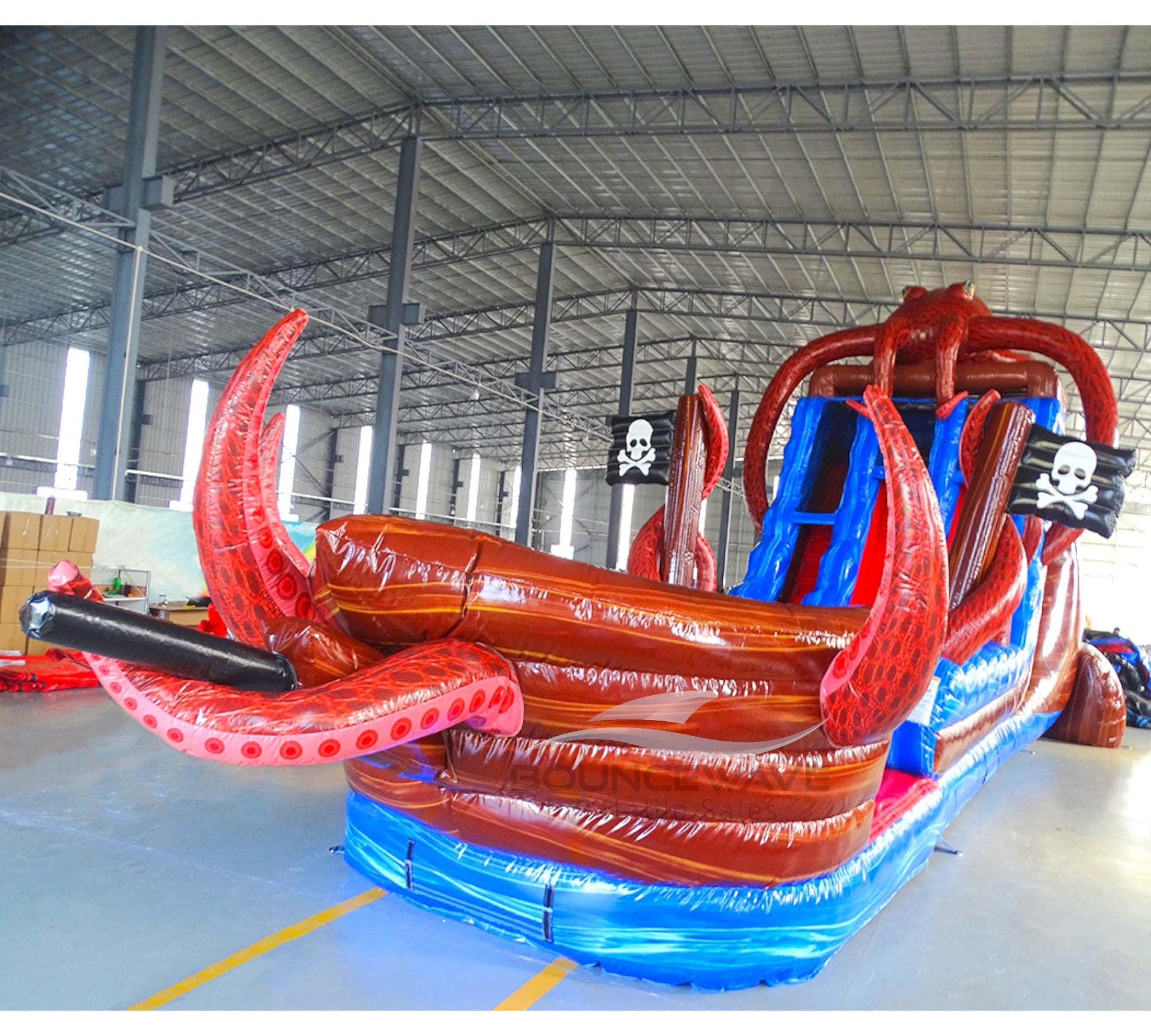 Side view of 20ft Tentacle Typhoon Hybrid DL water slide showing red, brown, and blue design, with large octopus tentacles wrapping around the structure. The slide features both open and enclosed sliding sections, displayed in an indoor facility with other inflatables visible.