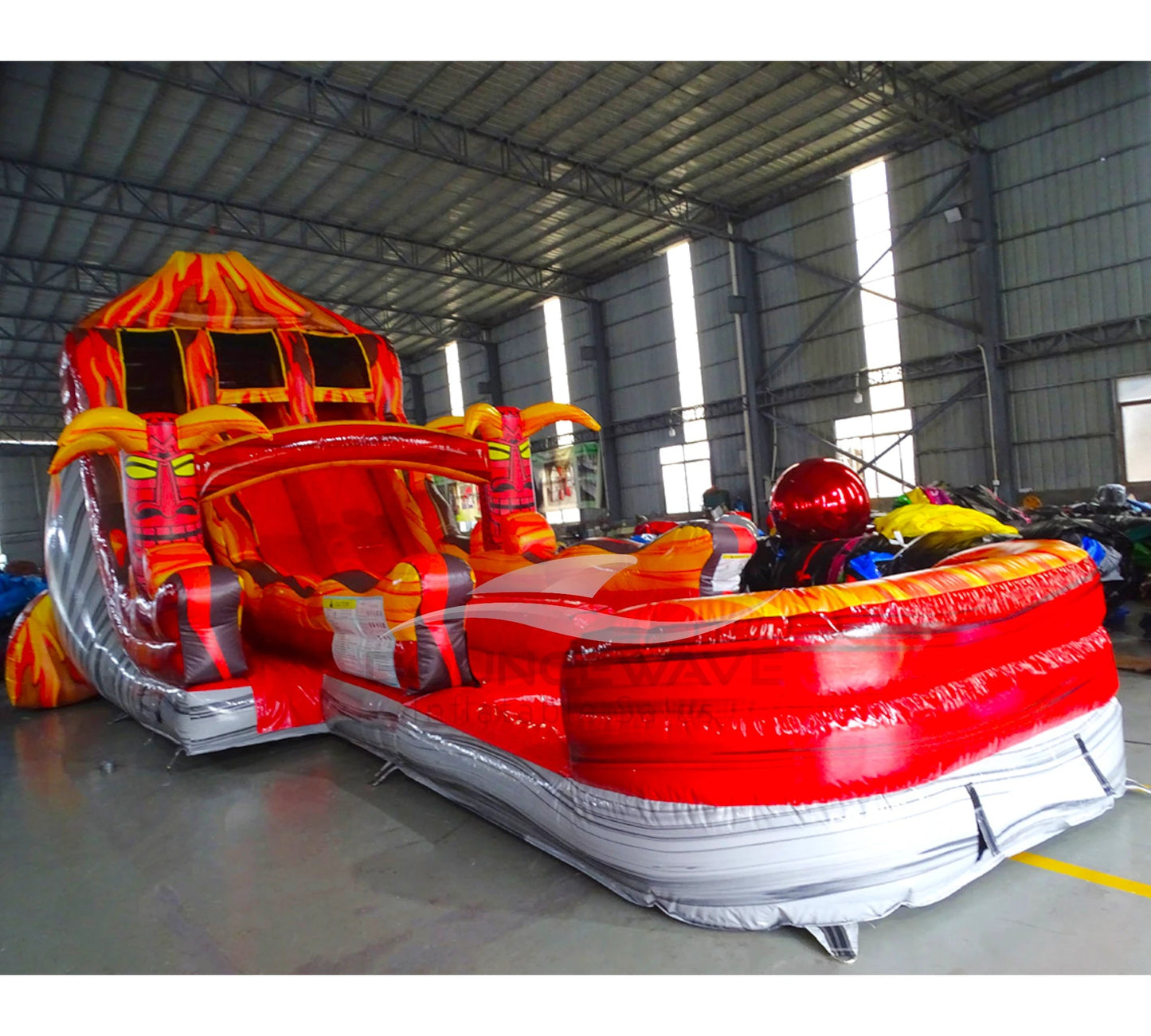 Side view of 20ft Tiki Volcano Hybrid DL water slide showing vibrant red, orange, and yellow colors resembling lava flow. The slide features a tiki face at the top, multiple sliding lanes, and a large splash pool area, displayed in an indoor facility with other inflatables visible.