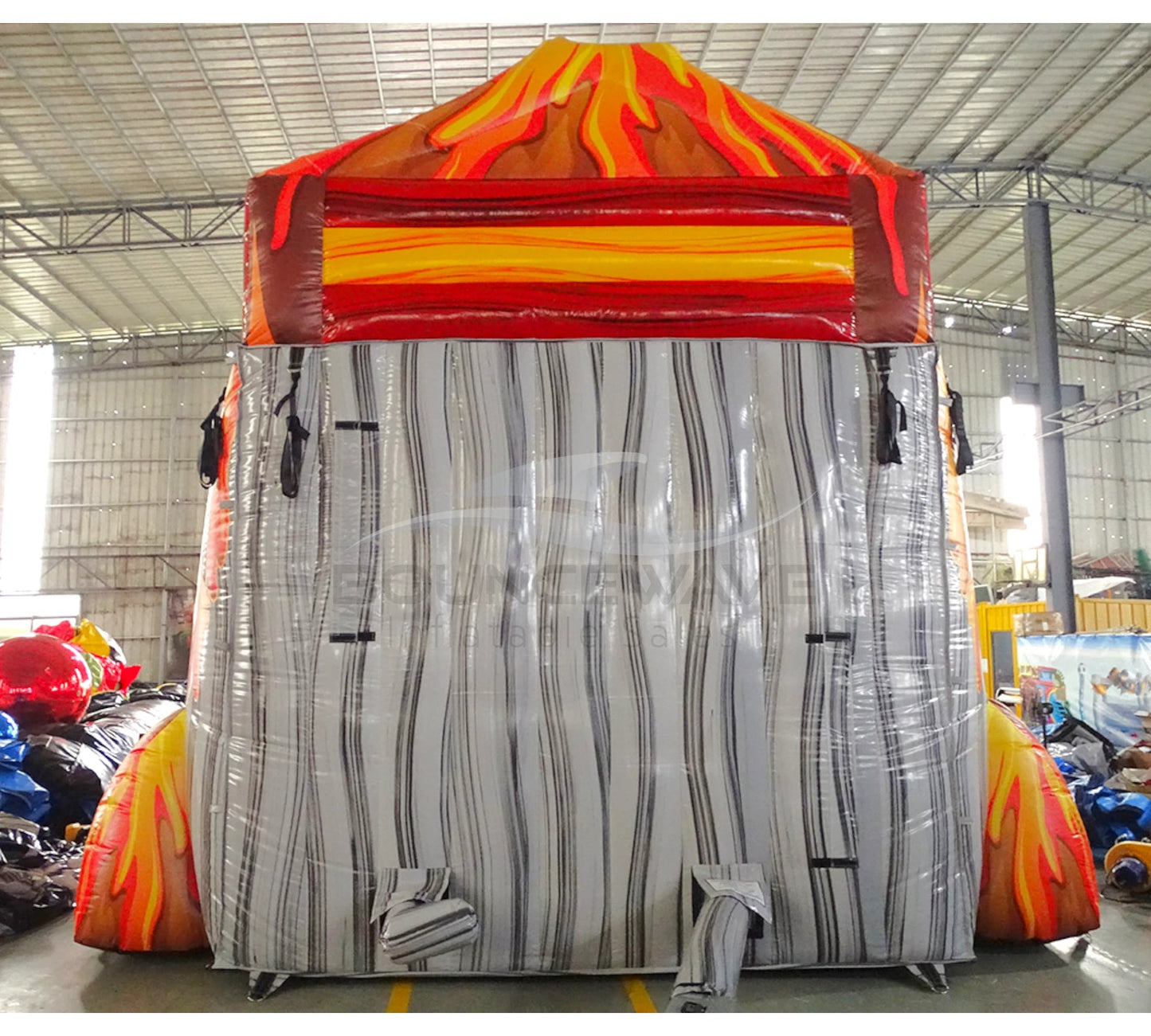 Large inflatable water slide with volcano theme, featuring red and orange colors, flame designs, and a tiki face at the top. The slide has multiple lanes, a spacious splash pool area, and combines open and enclosed sections, set up in an indoor warehouse.