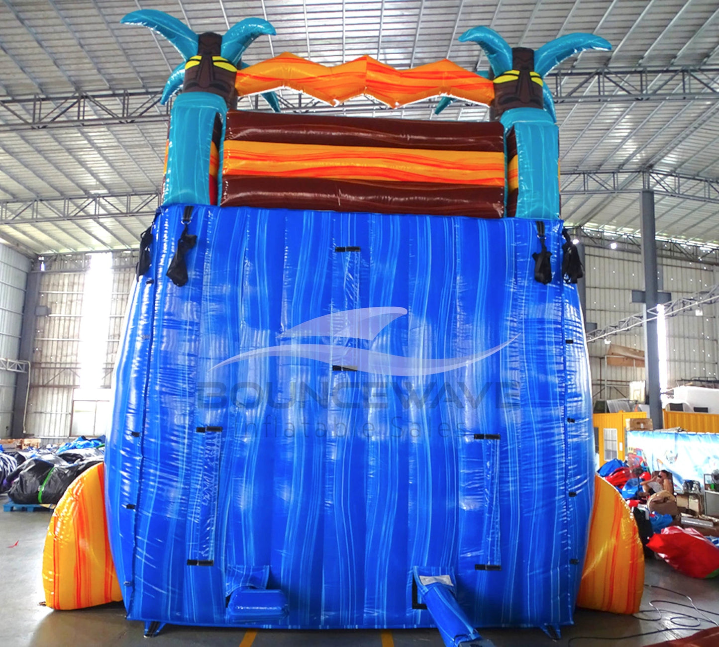 Large inflatable water slide with tropical theme, featuring vibrant blue, red, yellow, and orange colors, palm tree decorations, and a spacious splash pool area. The slide has multiple lanes, combines open and enclosed sections, and is set up in an indoor warehouse with metal roofing and other inflatable products visible in the background.