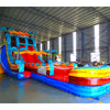 Large inflatable water slide with tropical theme, featuring vibrant blue, red, and yellow colors, palm tree decorations, and a spacious splash pool area. The slide has multiple lanes, combines open and enclosed sections, and is set up in an indoor warehouse with metal roofing visible.