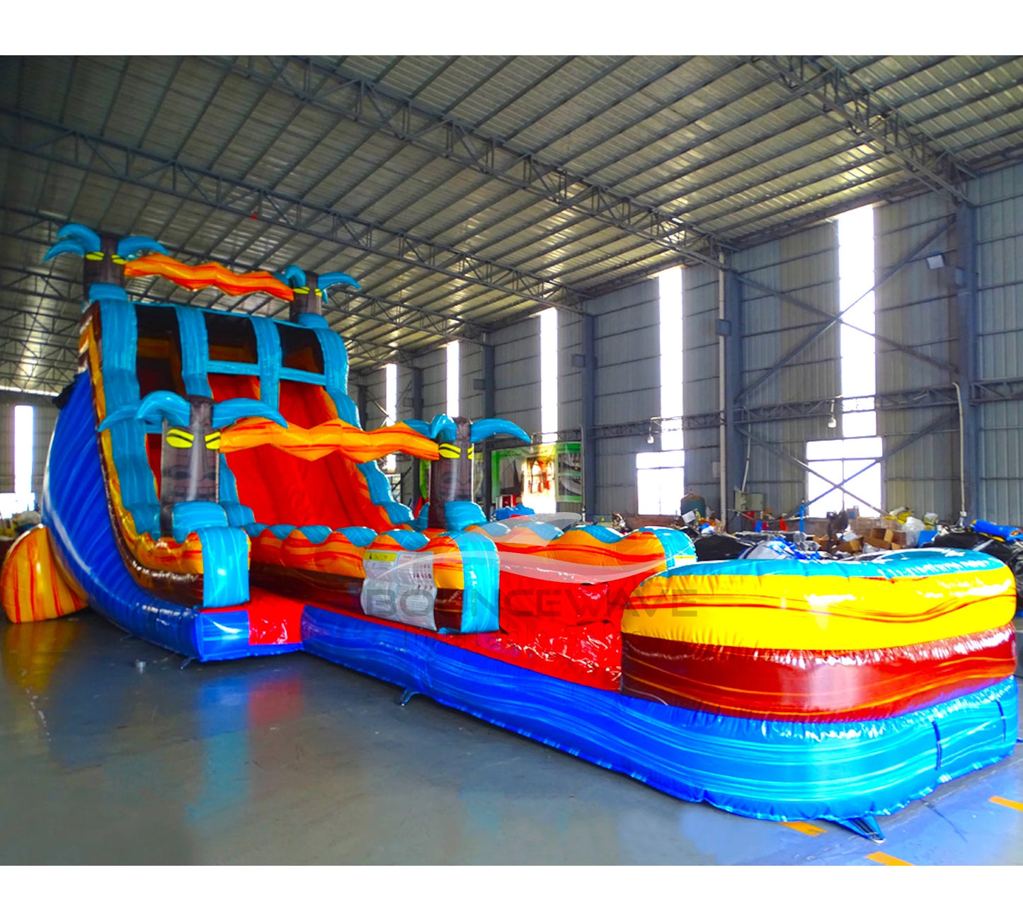 Large inflatable water slide with tropical theme, featuring vibrant blue, red, and yellow colors, palm tree decorations, and a spacious splash pool area. The slide has multiple lanes, combines open and enclosed sections, and is set up in an indoor warehouse with metal roofing visible.