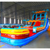 Side angle view of the 20ft Tropic Shock Hybrid DL inflatable water slide, showcasing its blue, red, and yellow sliding lanes, palm tree decorations, and spacious splash pool. The slide combines open and tube slide elements, set up in an indoor warehouse with other inflatables visible.