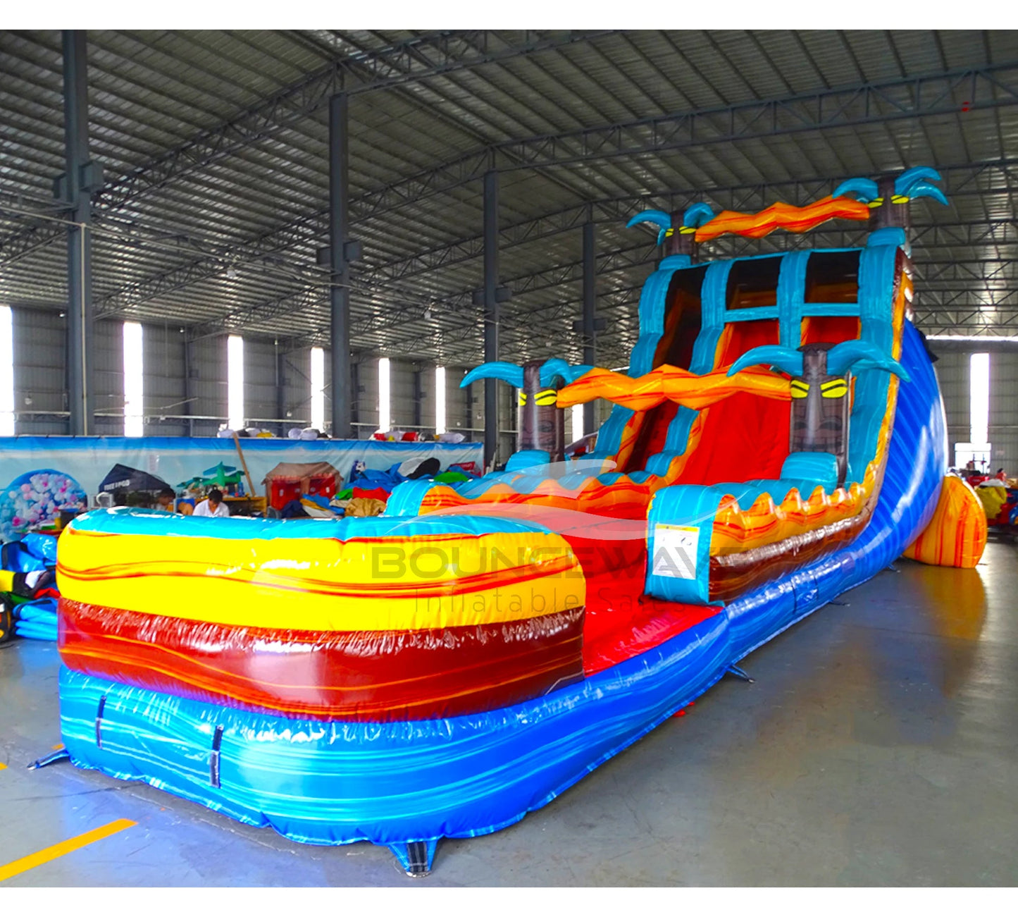 Side angle view of the 20ft Tropic Shock Hybrid DL inflatable water slide, showcasing its blue, red, and yellow sliding lanes, palm tree decorations, and spacious splash pool. The slide combines open and tube slide elements, set up in an indoor warehouse with other inflatables visible.