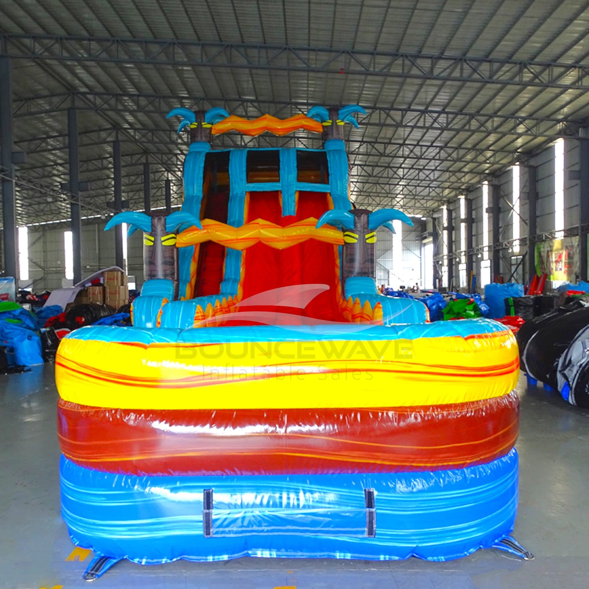 Front view of 20ft Tropic Shock Hybrid DL water slide showing bright blue, red, and yellow design, inflatable palm trees, and a large splash pool area with "BOUNCEWAVE" branding. The slide features both open and enclosed sliding sections, displayed in an indoor facility.