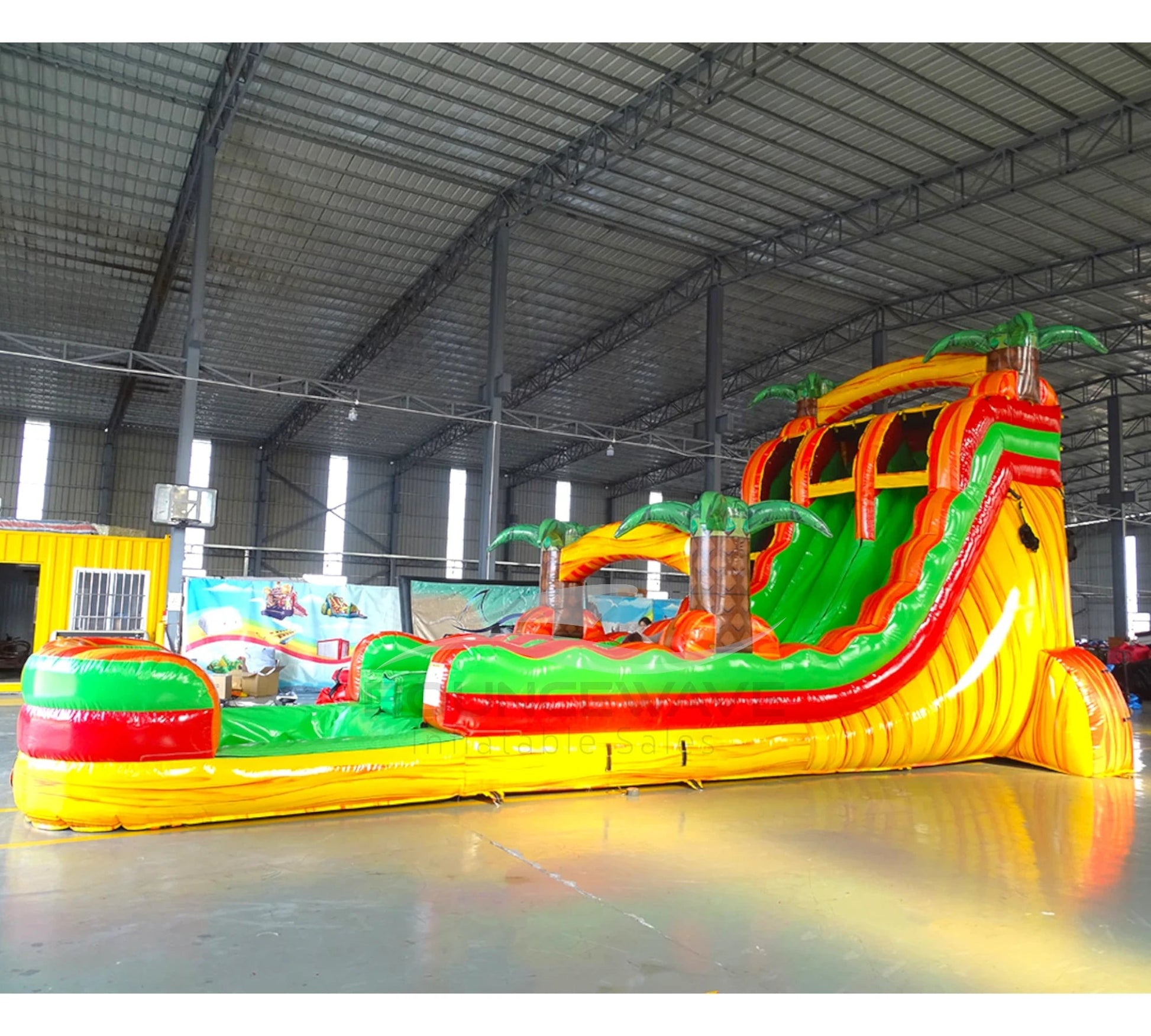 Angled view of the 20ft Tropical Inferno Hybrid DL inflatable water slide, showcasing its red, yellow, and green sliding lanes, palm tree decorations, and spacious splash pool. The slide combines open and tube slide elements, set up in an indoor warehouse with other equipment visible.