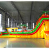 Side view of 20ft Tropical Inferno Hybrid DL water slide showing bright red, yellow, and green design, inflatable palm trees, and a large splash pool area. The slide features both open and enclosed sliding sections, displayed in an indoor facility with other inflatables visible.