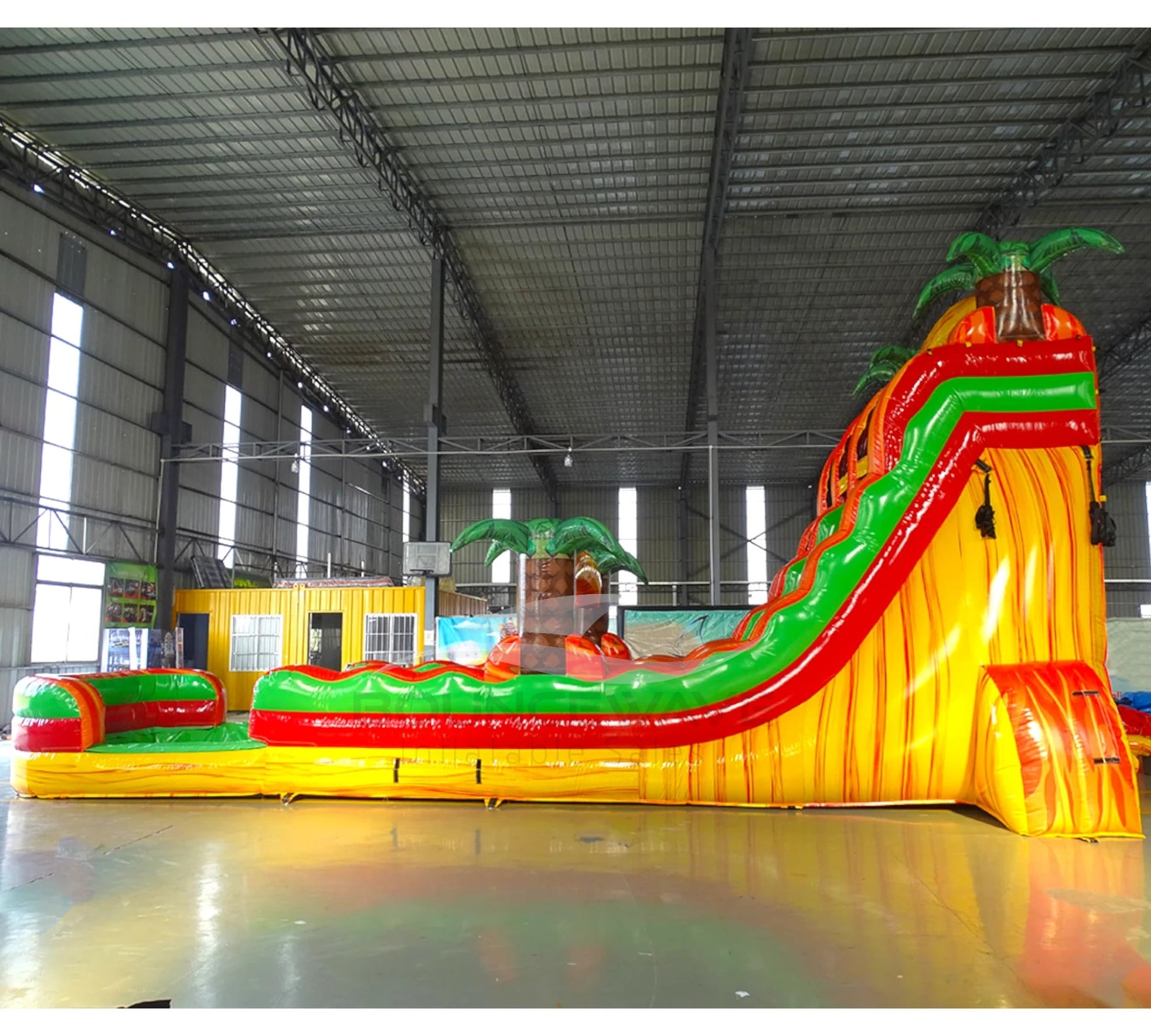 Side view of 20ft Tropical Inferno Hybrid DL water slide showing bright red, yellow, and green design, inflatable palm trees, and a large splash pool area. The slide features both open and enclosed sliding sections, displayed in an indoor facility with other inflatables visible.