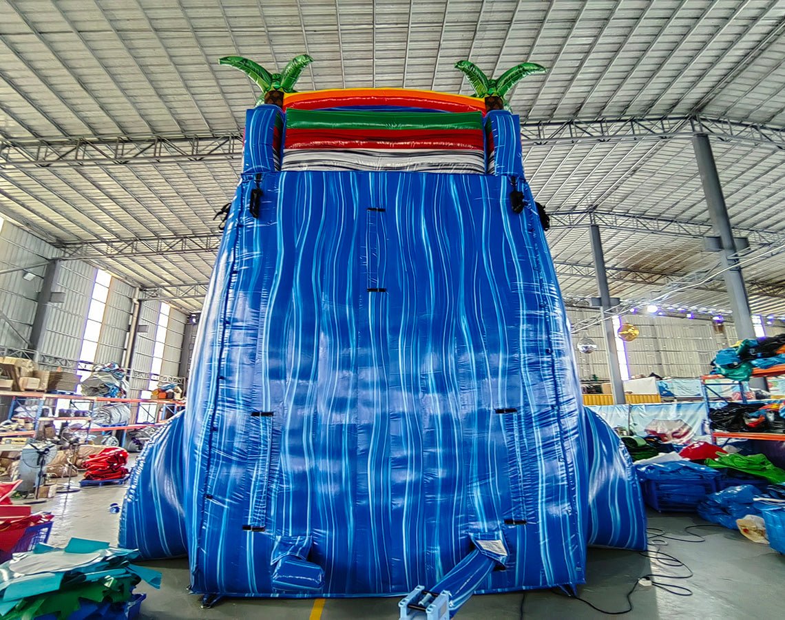 24ft Baja Splash DL 2pc Commercial Water Slide with Inflatable Slip & Slide featuring tropical palm trees, blue slide, and extended slip and slide in spacious warehouse