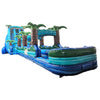 Large inflatable water slide with tropical theme, featuring bright blue colors, palm tree decorations, and a long slip and slide extension. The main slide has two lanes and a tall climbing wall. Set up in an indoor warehouse with metal roofing and other inflatable products visible in the background.