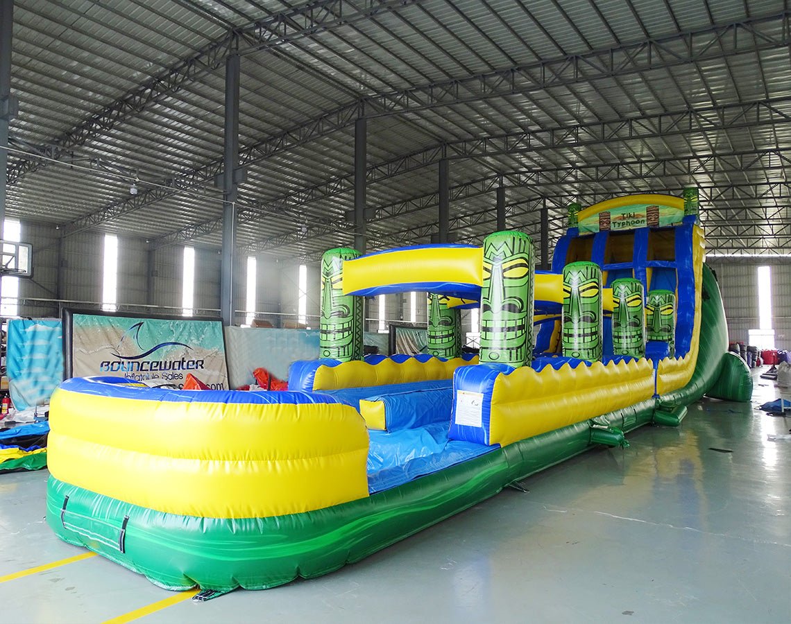 24ft Tiki Typhoon Dual Lane 2pc Commercial Water Slide with Inflatable Slip & Slide featuring tropical theme, tiki statues, and dual blue slides in warehouse setting
