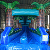 24ft Trop Wave DL 2pc Commercial Water Slide with Inflatable Slip & Slide featuring tropical palm trees, blue curved slide, and extended water play area in warehouse
