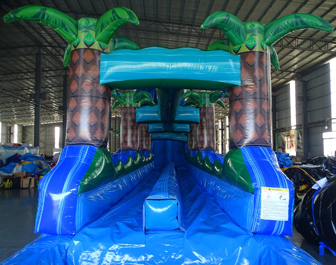 24ft Trop Wave DL 2pc Commercial Water Slide with Inflatable Slip & Slide featuring tropical palm trees, blue curved slide, and extended water play area in warehouse
