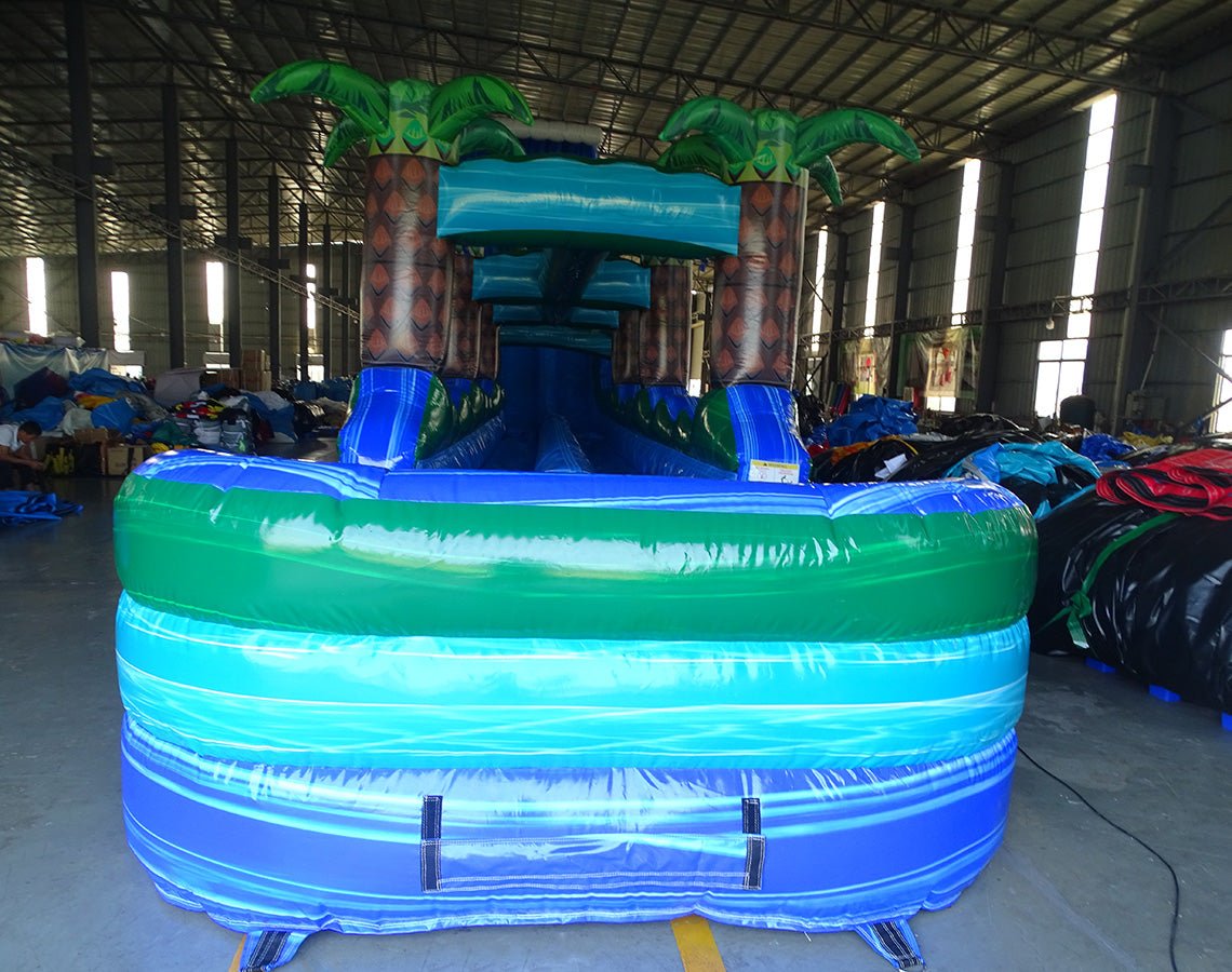 24ft Trop Wave DL 2pc Commercial Water Slide with Inflatable Slip & Slide featuring tropical palm trees, blue slide, and extended slip and slide in warehouse setting
