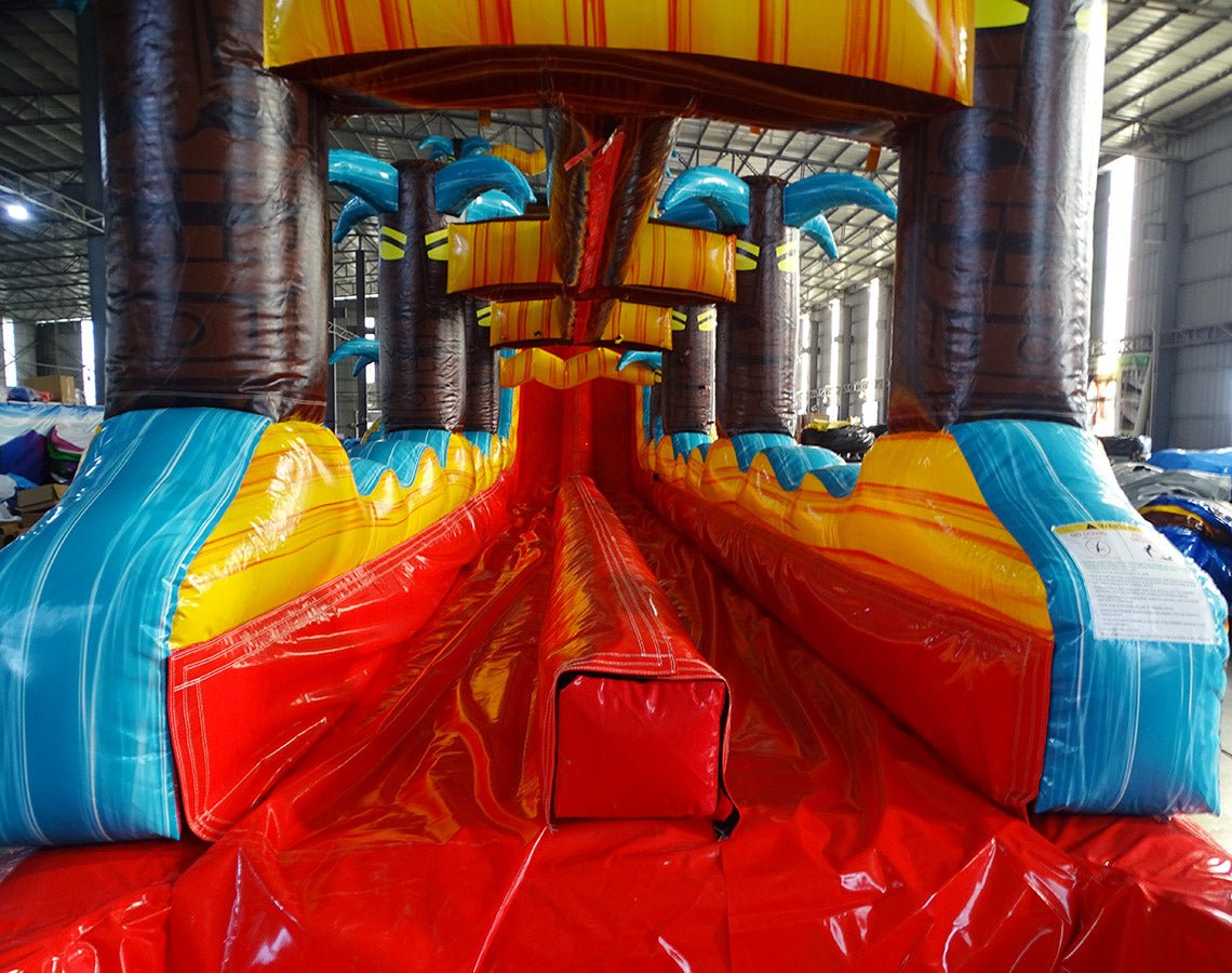 24ft Tropic Shock Dual Lane Commercial Water Slide with Inflatable Slip & Slide front view showcasing dual red slides, inflatable palm trees, and extended blue slip and slide
