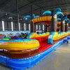 24ft Tropic Shock Dual Lane Commercial Water Slide with Inflatable Slip & Slide angled view highlighting palm tree decorations, blue dual slides, and yellow inflatable base in indoor facility
