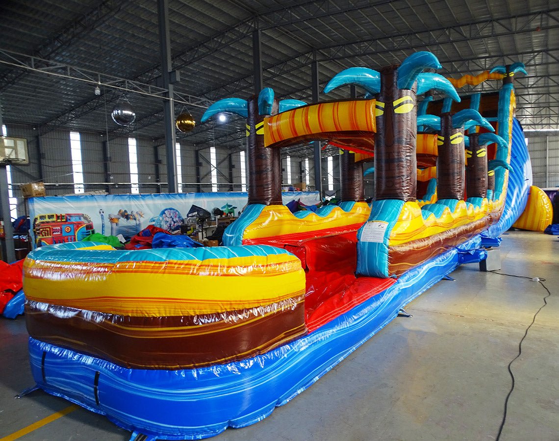 24ft Tropic Shock Dual Lane Commercial Water Slide with Inflatable Slip & Slide angled view highlighting palm tree decorations, blue dual slides, and yellow inflatable base in indoor facility
