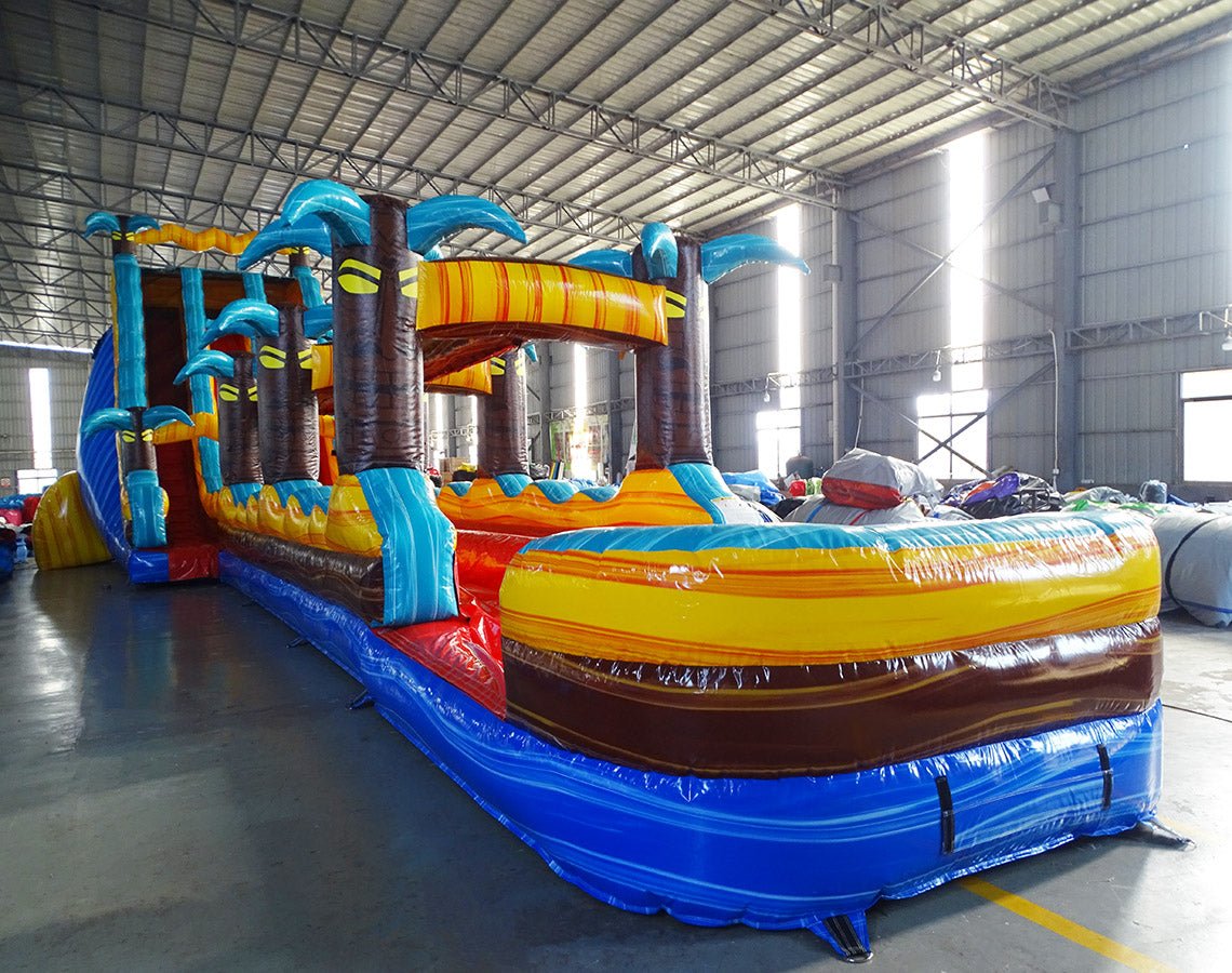 24ft Tropic Shock Dual Lane Commercial Water Slide with Inflatable Slip & Slide side view showcasing vibrant colors, inflatable palm trees, and long slip and slide attachment
