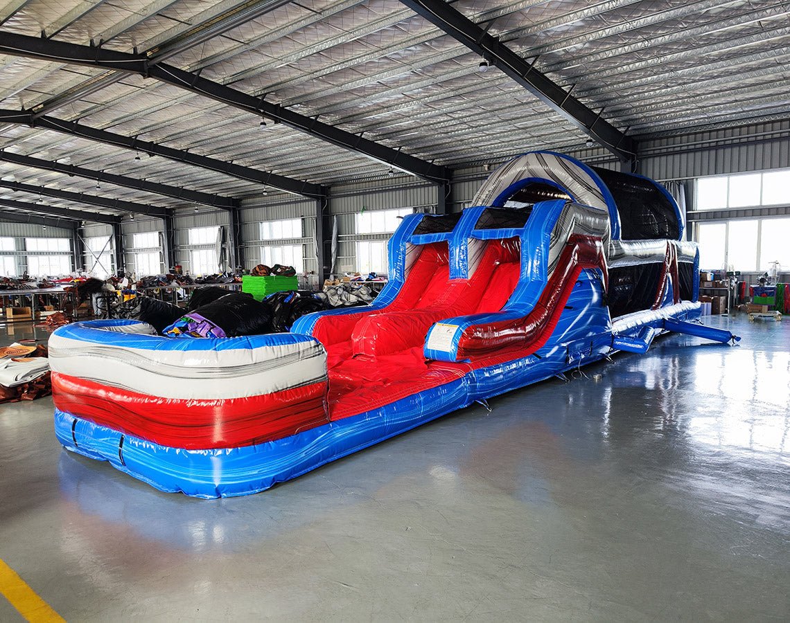 Side view of 46ft Baja Commercial Hybrid Inflatable Obstacle Course in a large indoor warehouse. The inflatable features a red, white, and blue color scheme with multiple obstacles including slides, tunnels, and climbing walls. The course stretches across the warehouse floor, showcasing its length. Metal roofing, support beams, and windows are visible in the background.