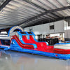 Front angled view of 46ft Baja Commercial Hybrid Inflatable Obstacle Course set up in a spacious warehouse. The inflatable structure has a patriotic red, white, and blue design with various obstacles visible, including slides, tunnels, and climbing sections. The warehouse interior shows metal roofing, support beams, and a white partition wall in the background, providing context for the obstacle course's size and commercial setting.