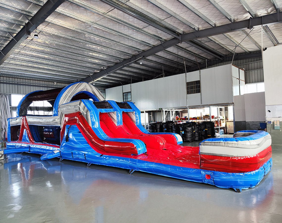 Front angled view of 46ft Baja Commercial Hybrid Inflatable Obstacle Course set up in a spacious warehouse. The inflatable structure has a patriotic red, white, and blue design with various obstacles visible, including slides, tunnels, and climbing sections. The warehouse interior shows metal roofing, support beams, and a white partition wall in the background, providing context for the obstacle course's size and commercial setting.