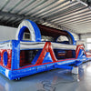 Front angled view of 46ft Baja Commercial Hybrid Inflatable Obstacle Course in a warehouse. Shows entrance with inflatable archway, followed by a series of obstacles in red, white, and blue. Warehouse interior visible with metal ceiling and other equipment, providing scale context.