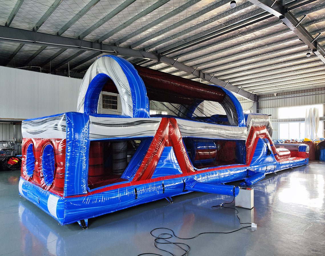 Front angled view of 46ft Baja Commercial Hybrid Inflatable Obstacle Course in a warehouse. Shows entrance with inflatable archway, followed by a series of obstacles in red, white, and blue. Warehouse interior visible with metal ceiling and other equipment, providing scale context.