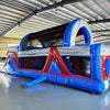 Wide-angle view of 46ft Baja Commercial Hybrid Inflatable Obstacle Course in a large indoor warehouse. Red, white, and blue inflatable structure with various obstacles visible. Metal roofing, concrete flooring, and other inflatable products in background, demonstrating its size and commercial setting.