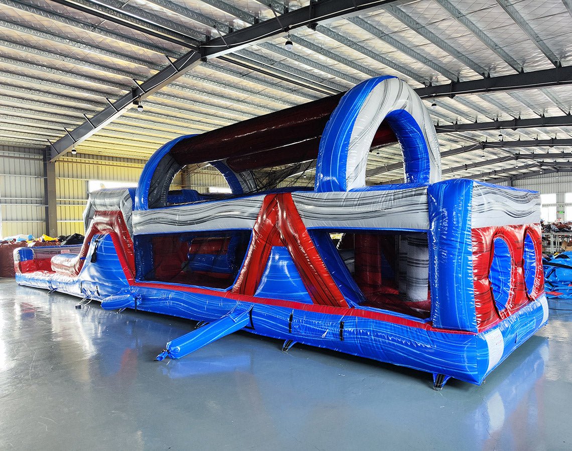 Wide-angle view of 46ft Baja Commercial Hybrid Inflatable Obstacle Course in a large indoor warehouse. Red, white, and blue inflatable structure with various obstacles visible. Metal roofing, concrete flooring, and other inflatable products in background, demonstrating its size and commercial setting.