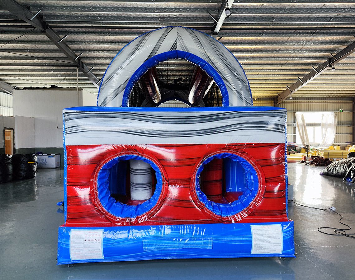 Close-up rear view of 46ft Baja Commercial Hybrid Inflatable Obstacle Course entrance. Features inflatable archway with "START" text, followed by initial obstacles. Red, white, and blue color scheme prominent. Set in a warehouse with metal roofing and concrete floor visible.