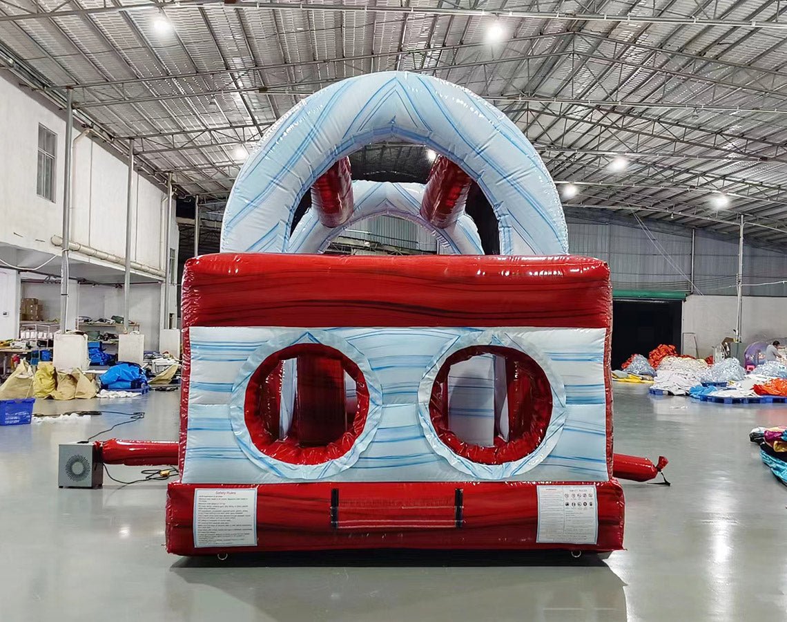 Rear view of 46ft Crimson Bay Commercial Hybrid Inflatable Obstacle Course set up in a large indoor warehouse. The inflatable features a red base with blue accents, multiple climbing walls, slides, and obstacles. Metal roofing and various other inflatable products are visible in the background.