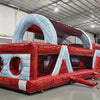 Three-quarter view of 46ft Crimson Bay Commercial Hybrid Inflatable Obstacle Course in a spacious warehouse. The inflatable displays a red and blue design with multiple obstacles, including climbing walls, tunnels, and slides. Other inflatable products and warehouse materials are visible in the background, demonstrating its size and commercial setting.