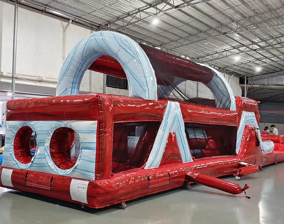 Three-quarter view of 46ft Crimson Bay Commercial Hybrid Inflatable Obstacle Course in a spacious warehouse. The inflatable displays a red and blue design with multiple obstacles, including climbing walls, tunnels, and slides. Other inflatable products and warehouse materials are visible in the background, demonstrating its size and commercial setting.