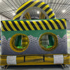 Rear view of 46ft Hazardous Commercial Hybrid Inflatable Obstacle Course showing the entrance area. The inflatable features yellow and black hazard stripes, circular openings, and a diamond plate pattern. Safety instructions are visible on the base. Set in a warehouse with concrete flooring and various other inflatable products in the background.