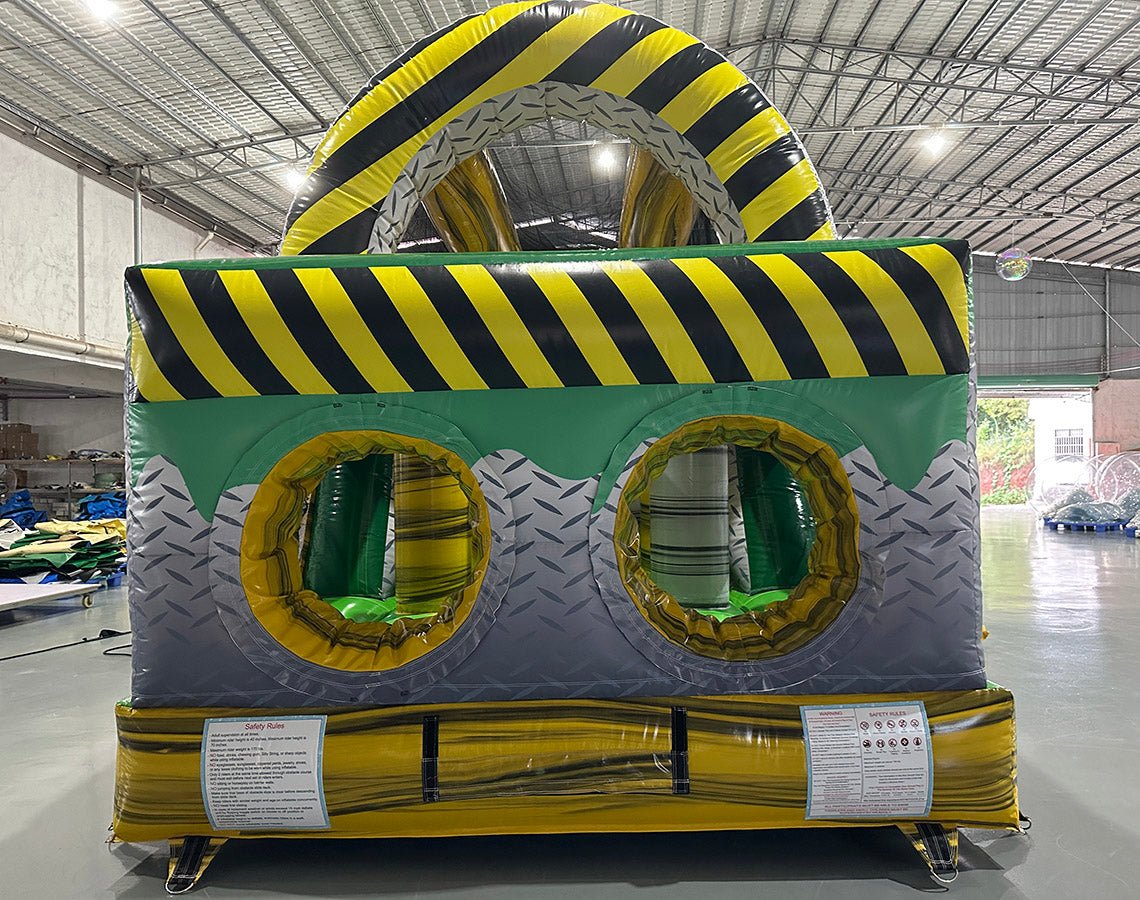 Rear view of 46ft Hazardous Commercial Hybrid Inflatable Obstacle Course showing the entrance area. The inflatable features yellow and black hazard stripes, circular openings, and a diamond plate pattern. Safety instructions are visible on the base. Set in a warehouse with concrete flooring and various other inflatable products in the background.